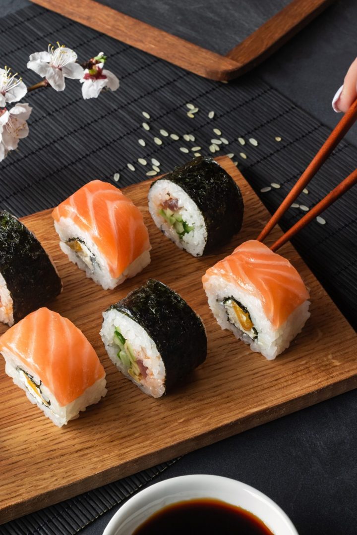 Set of sushi and maki rolls, hand with chopsticks and branch of white flowers on stone table